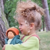 Petite fille portant dans ses bras une poupée Nanchen avec robe rayée blanc et vert, veste en laine verte à capuche et bonnet de laine orange.